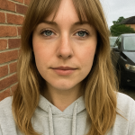 Woman with medium-blonde hair and bangs taking a casual selfie against a red brick wall, light gray hoodie, parked car in background.