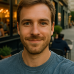 Man with green eyes and light brown-blond hair taking an outdoor café selfie at dusk, casual blue T-shirt and trimmed scruff.