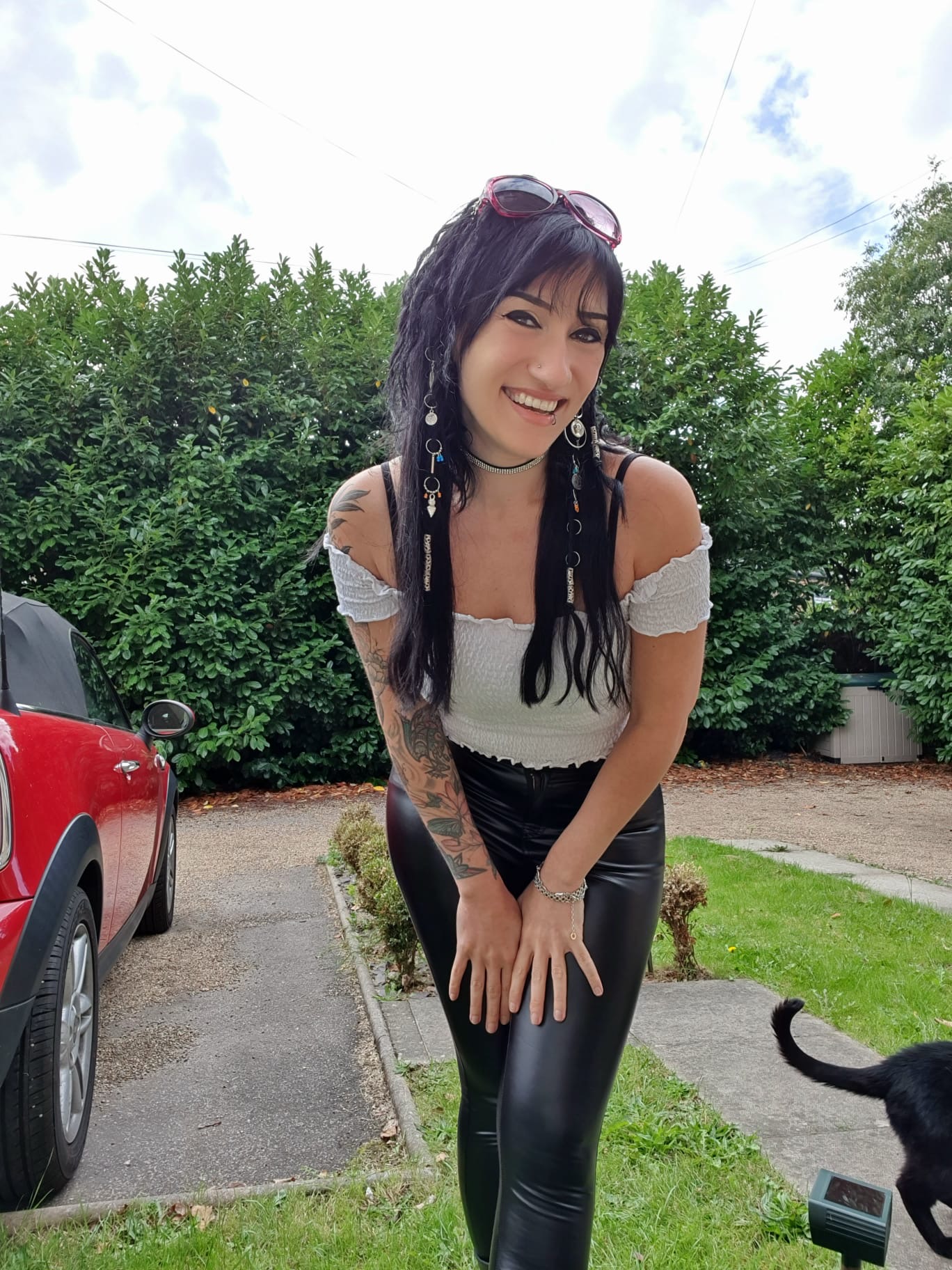 Athena Raven smiling and leaning towards the camera outdoors, wearing an off-the-shoulder top and black pants.