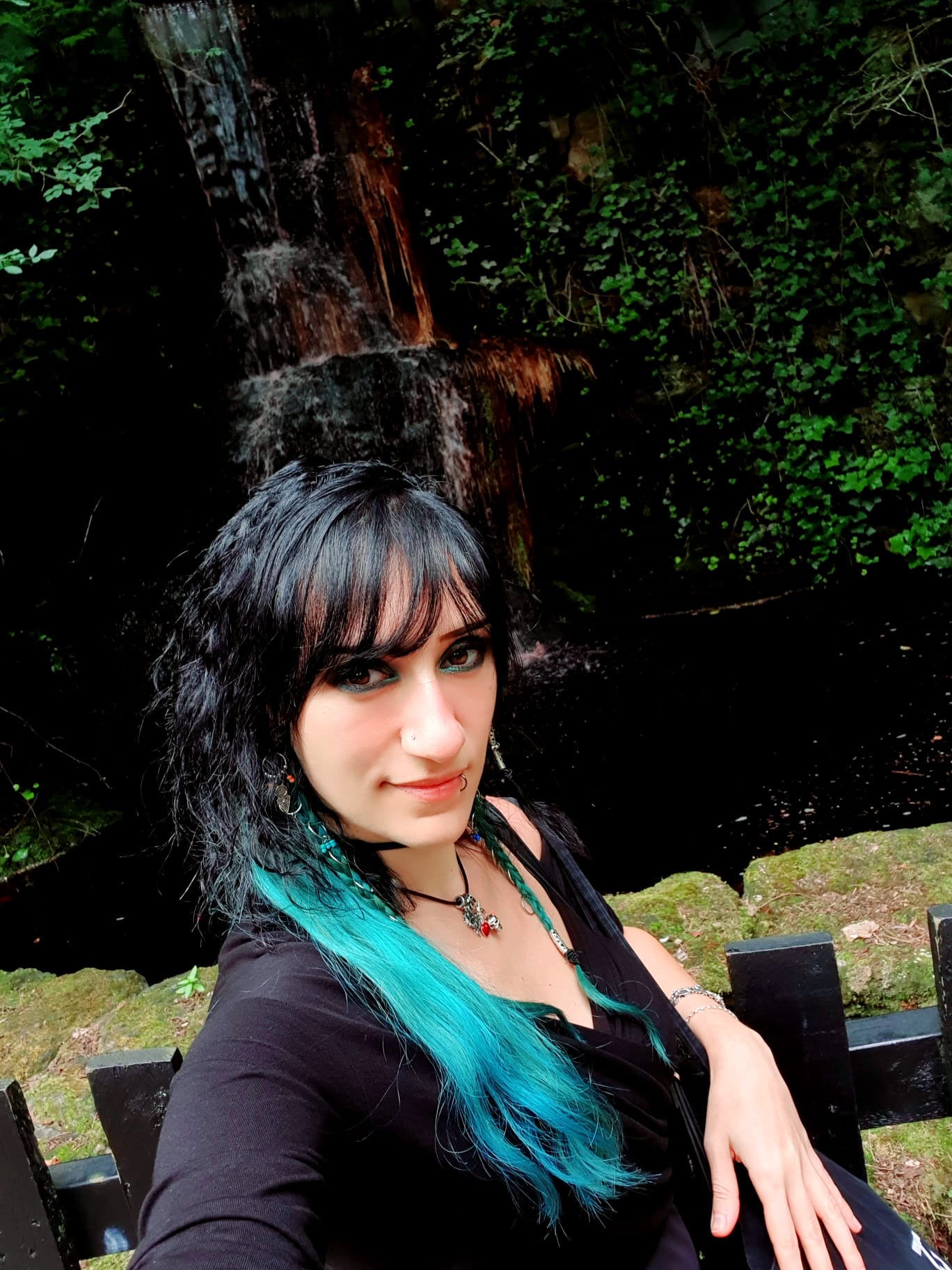 Athena Raven sitting on a park bench next to a cello, with long dark hair featuring turquoise highlights.