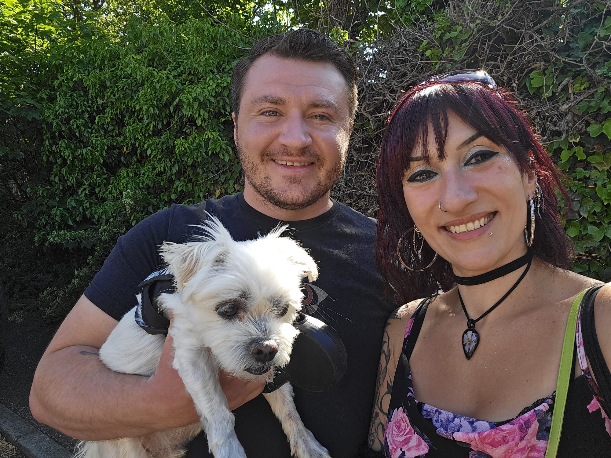 Joseph Alai, a man with a beard, holding a small white dog, Koko Bean, and smiling next to Athena Raven outdoors.